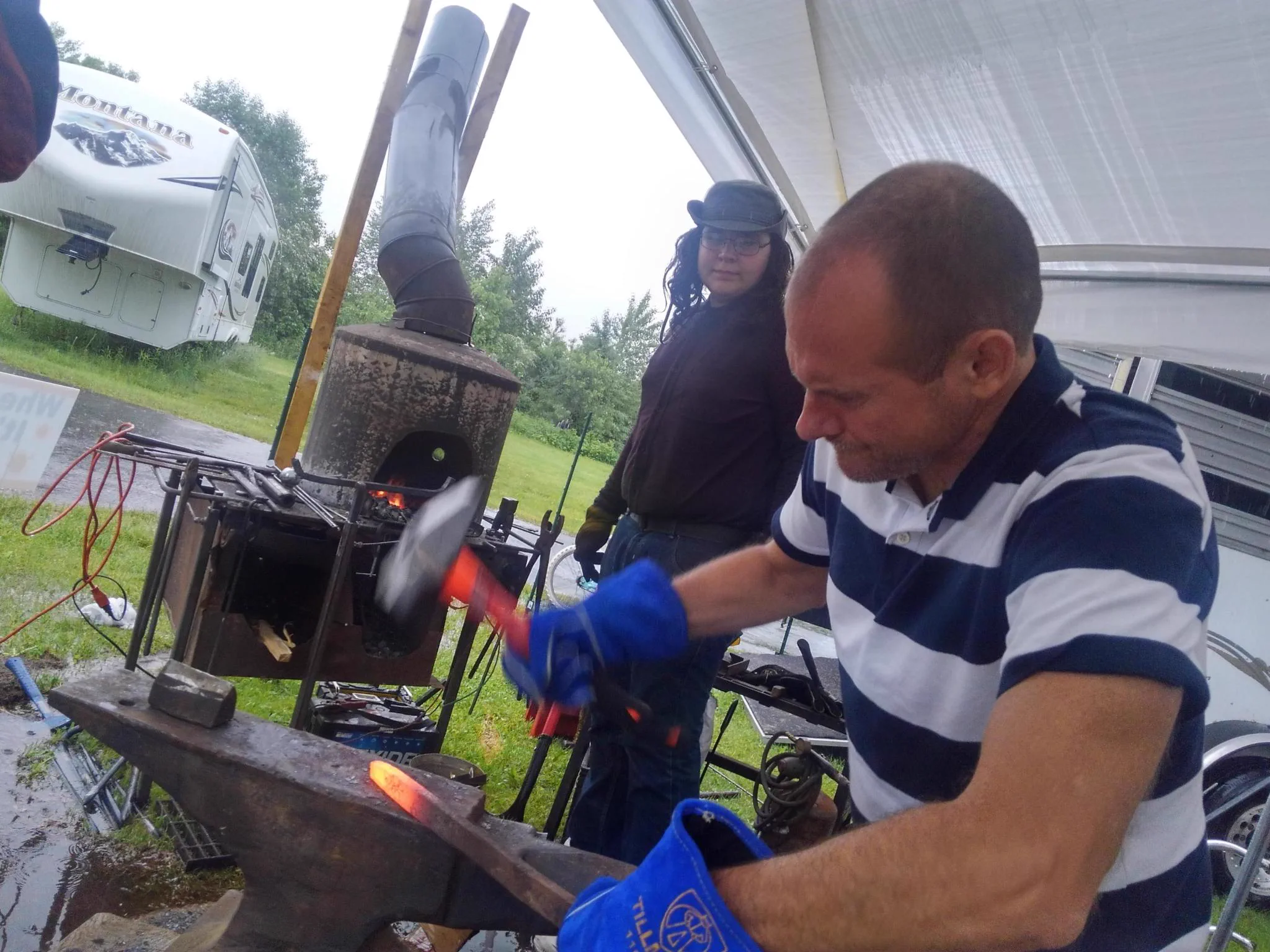 Etienne de la Boetie, a volunteer blacksmith mentor, holding glowing hot iron with tongs on an anvil, with a coal forge behind him